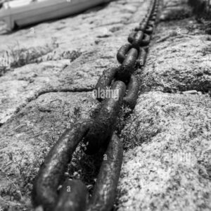 Close-up of a rusty iron chain on a stone dock with boats blurred in the background, black and white photo.