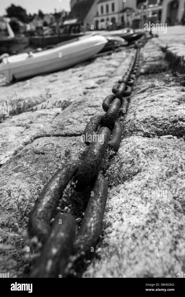 Close-up of a rusty iron chain on a stone dock with boats blurred in the background, black and white photo.