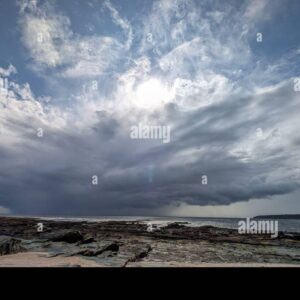 Dark storm clouds forming over the ocean with sunlight breaking through above a rocky shoreline.