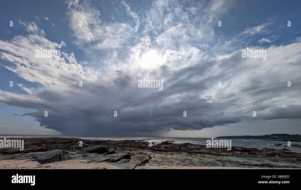 Dark storm clouds forming over the ocean with sunlight breaking through above a rocky shoreline.