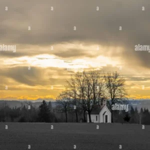 A small white chapel with bare trees at sunset under a dramatic cloudy sky.