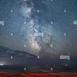 Night sky with the Milky Way over the ocean, with a rocky coastline illuminated in red light in the foreground.