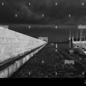 Black and white night photo of a harbor walkway with yachts moored on the side.