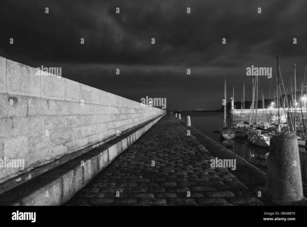 Black and white night photo of a harbor walkway with yachts moored on the side.