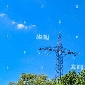 High voltage electricity pylon with power lines above green trees against clear blue sky.