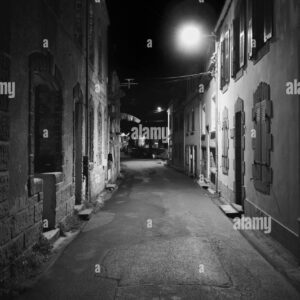 Deserted narrow street at night illuminated by street lamps in black and white.