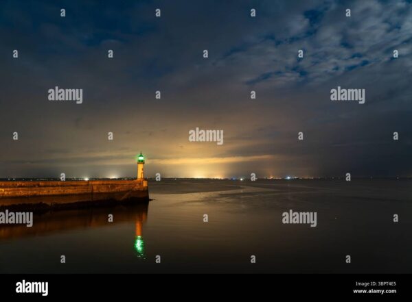 Lighthouse with green light glowing at night by the sea under dramatic sky.